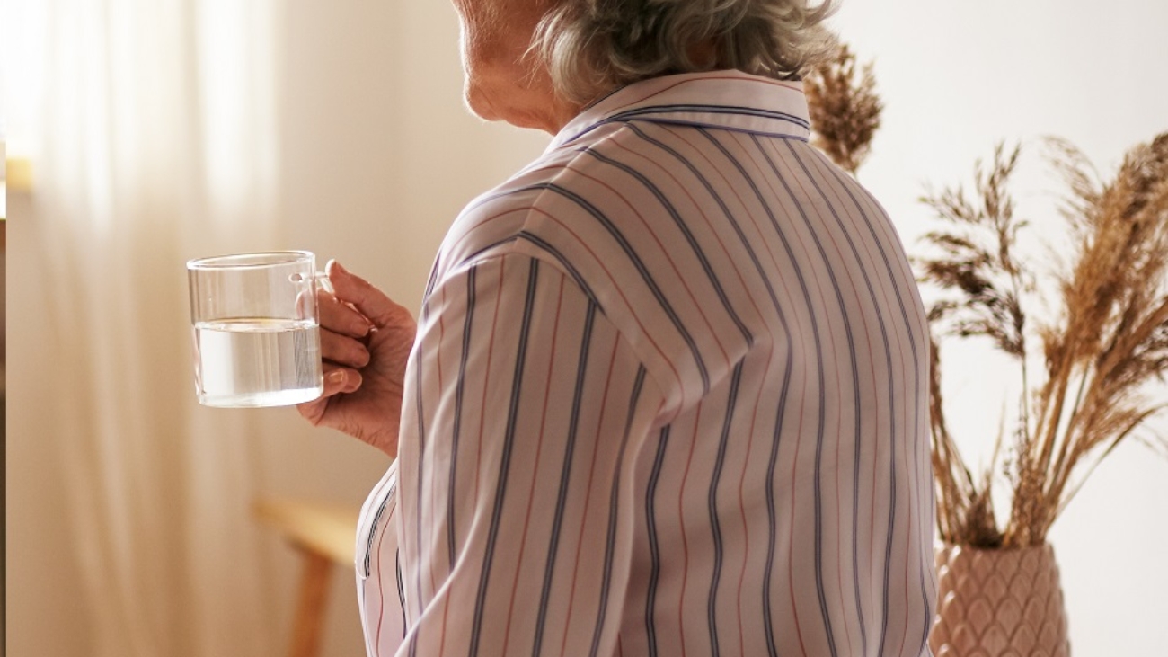 Rear view of senior sixty year old woman with gray hair holding mug washing down sleeping pill, suffering from insomnia. Elderly retired female taking medicine with water, sitting in bedroom