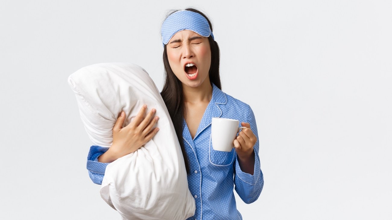Morning lifestyle, breakfast and people concept. Girl with insomnia in sleeping mask and pajamas, hugging pillow, drinking coffee and yawning, trying wake-up, white background.